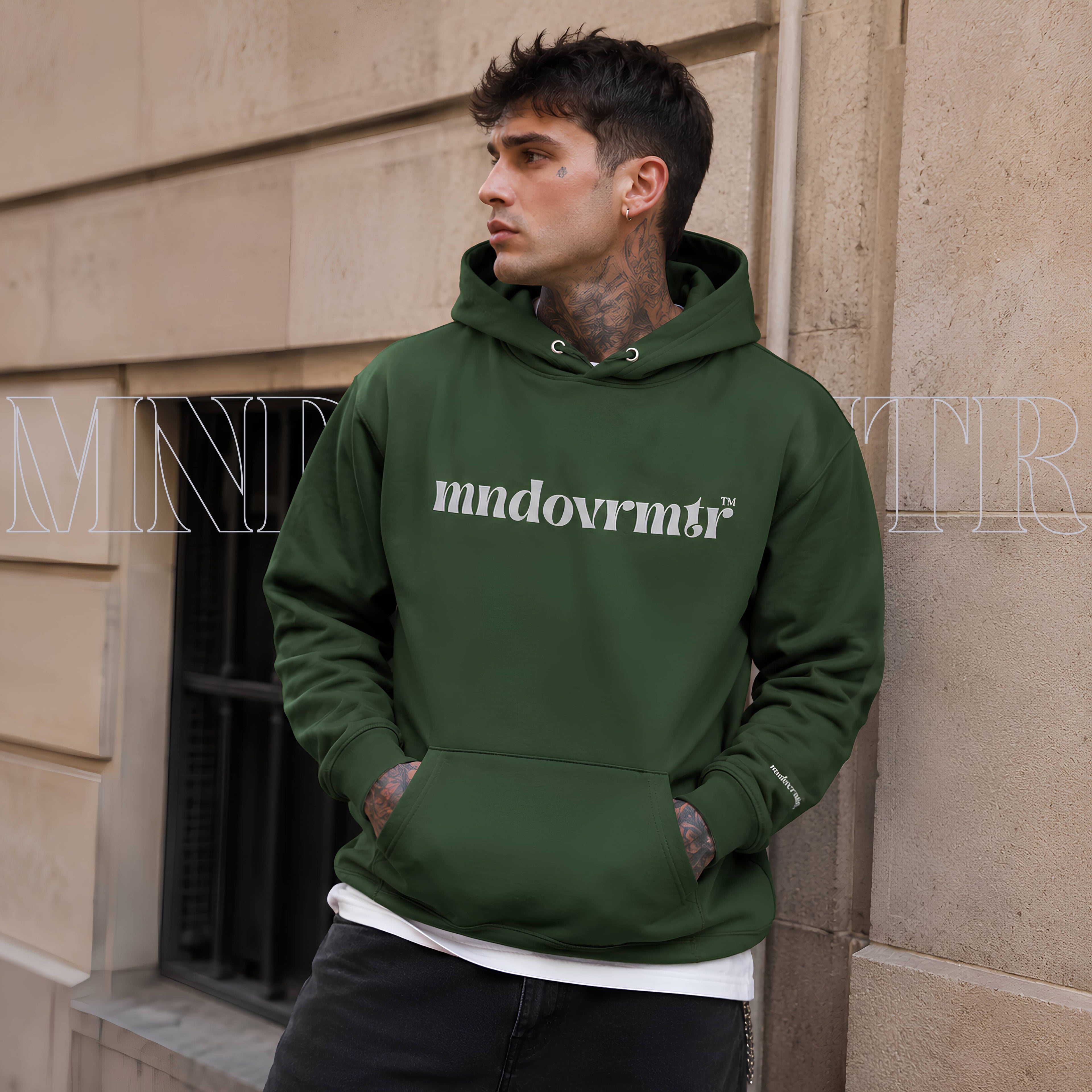 A gorgeous man, casually leaning on a wall. He is wearing a forest green, cotton, fleece, oversized, unisex hoodie, with a gray 'mndovrmtr' text across the chest