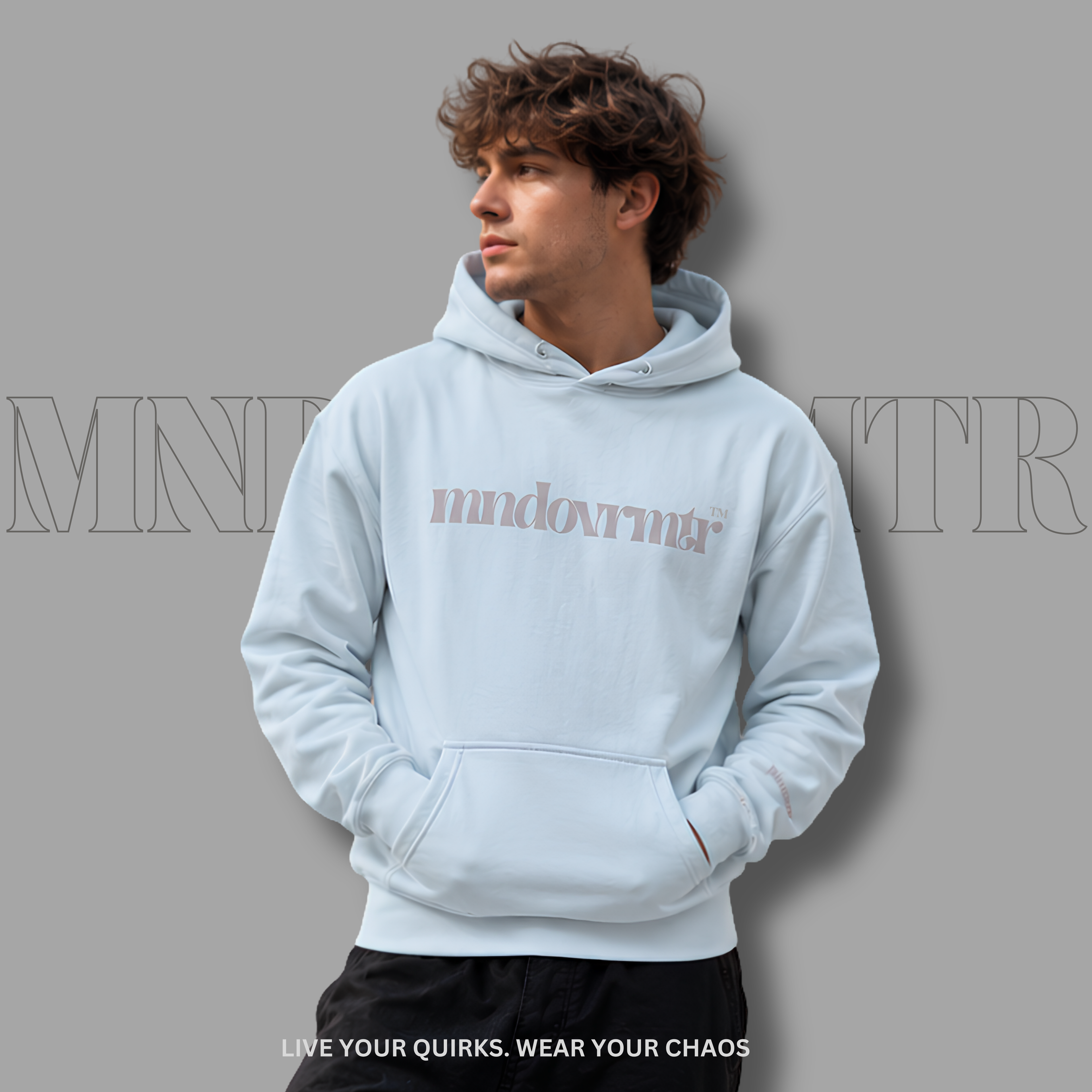 A man wearing a light blue, fleece, oversized, front pocket, pullover hoodie with 'mndovrmtr' printed in gray text across the chest, posed against a solid gray background with a subtle drop shadow.
