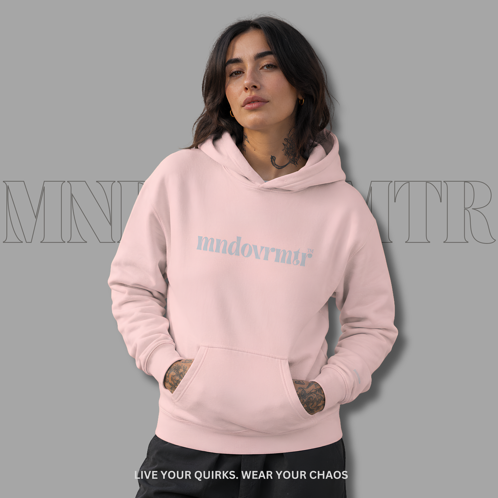 A woman wearing a light pink, fleece, oversized, cotton, with front pocket, pullover hoodie with the word 'mndovrmtr' printed in a gray script across the chest, posed against a solid gray background.