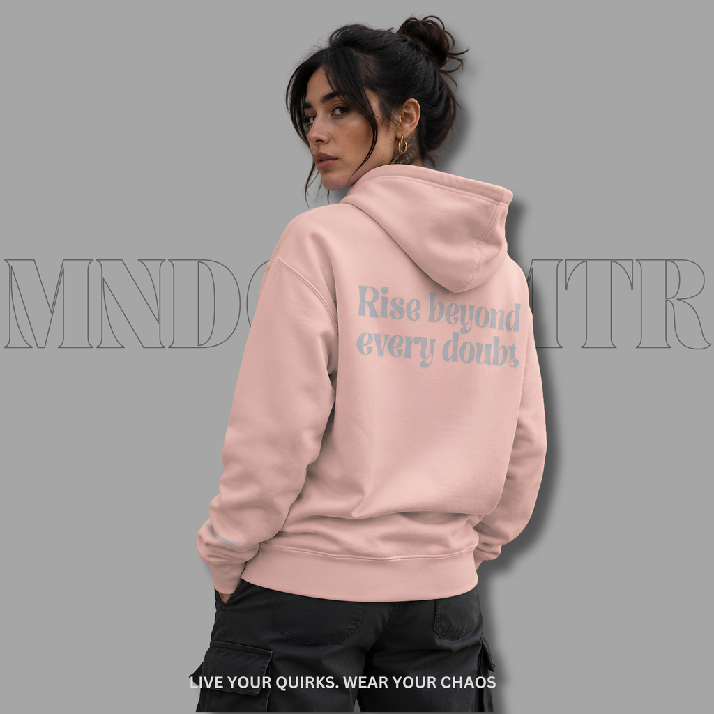 A woman wearing a light pink, oversized, fleece, cotton, hoodie with the text 'Rise beyond every doubt' printed in gray on the back, shown from behind against a solid gray background.