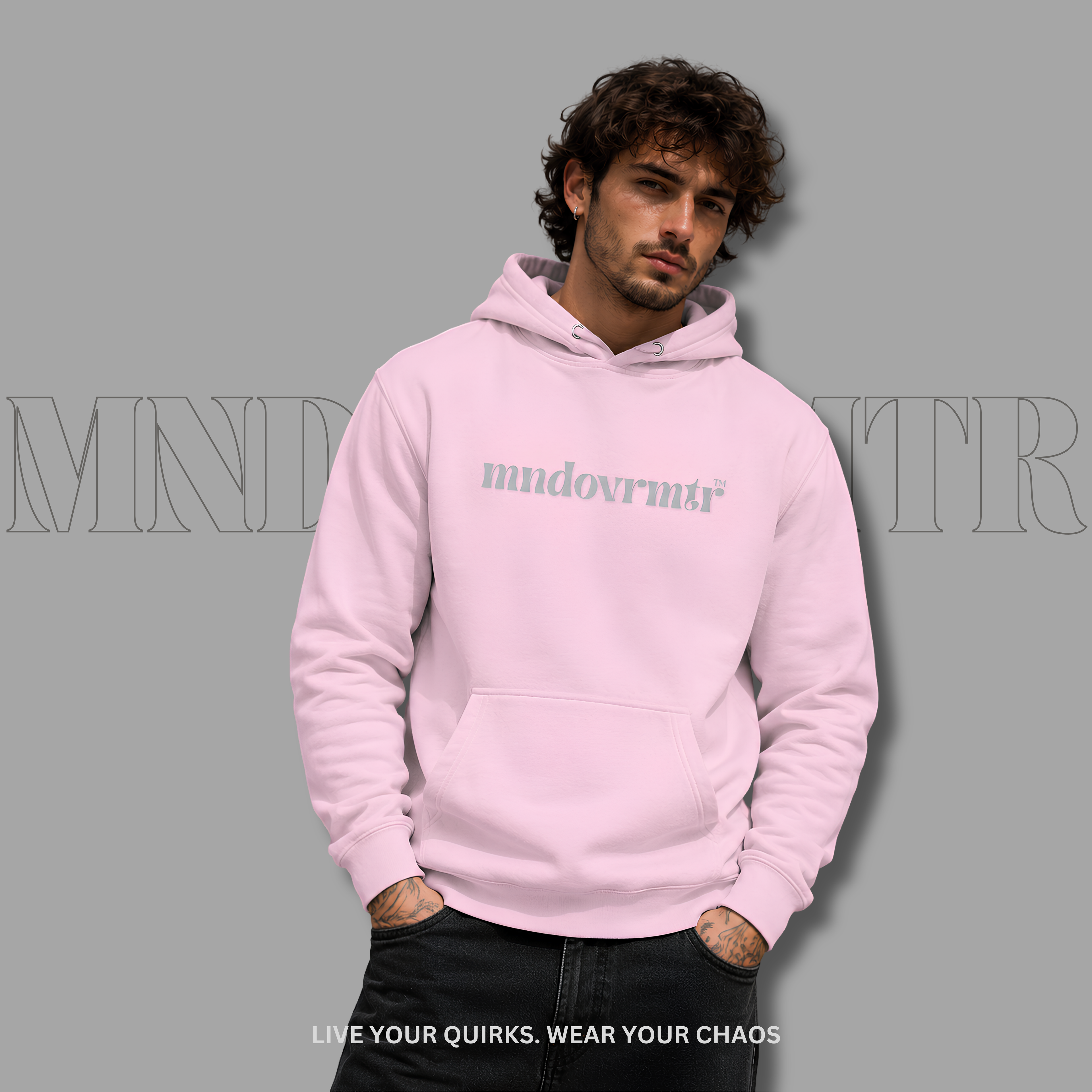 A man wearing a light pink, fleece, oversized, cotton, front pocket, hoodie with the text 'mndovrmtr' printed on the chest, posed against a solid gray background with a faint brand watermark.