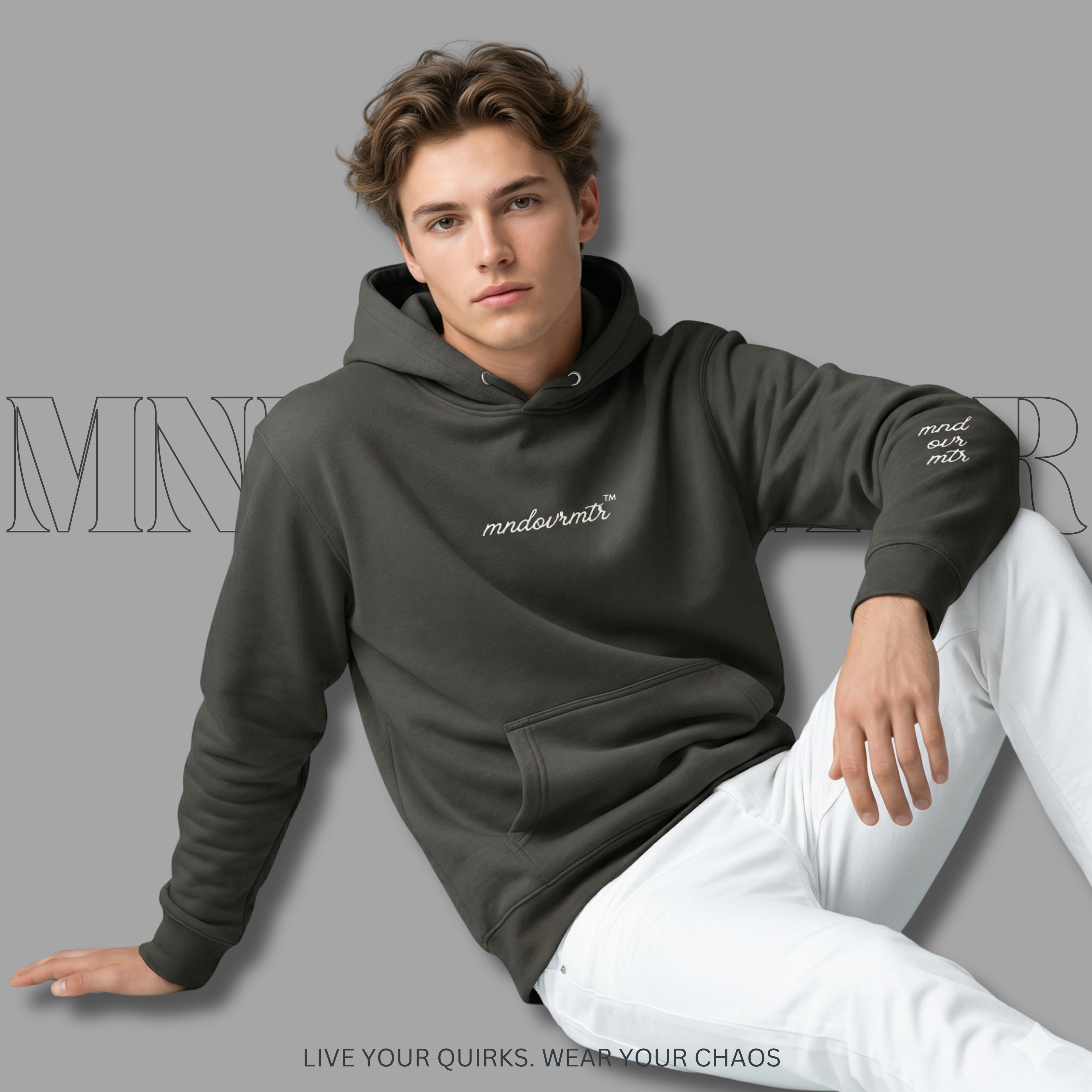MNDOVRMTR Man wearing a dark gray hoodie with text, sitting on a light gray couch against a gray background.
